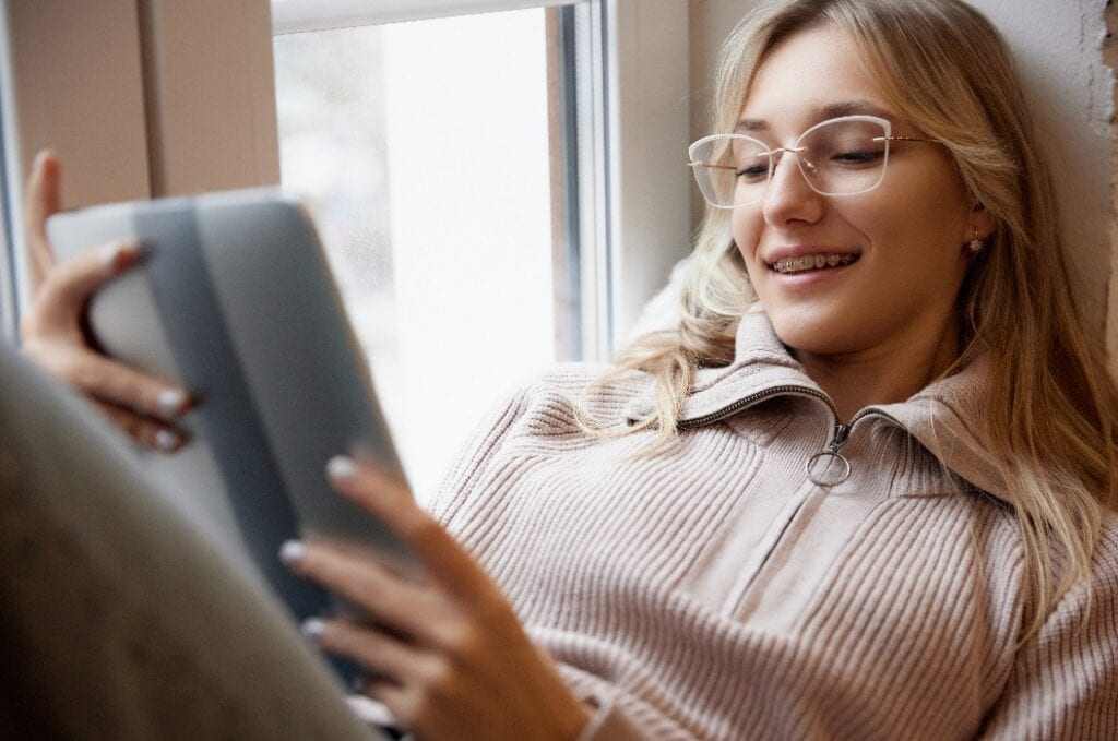 Junge Frau mit Zahnspange, die einen beigen Pullover und eine Brille trägt und lächelnd ein Tablet am Fenster liest. Lächelnder Patient.