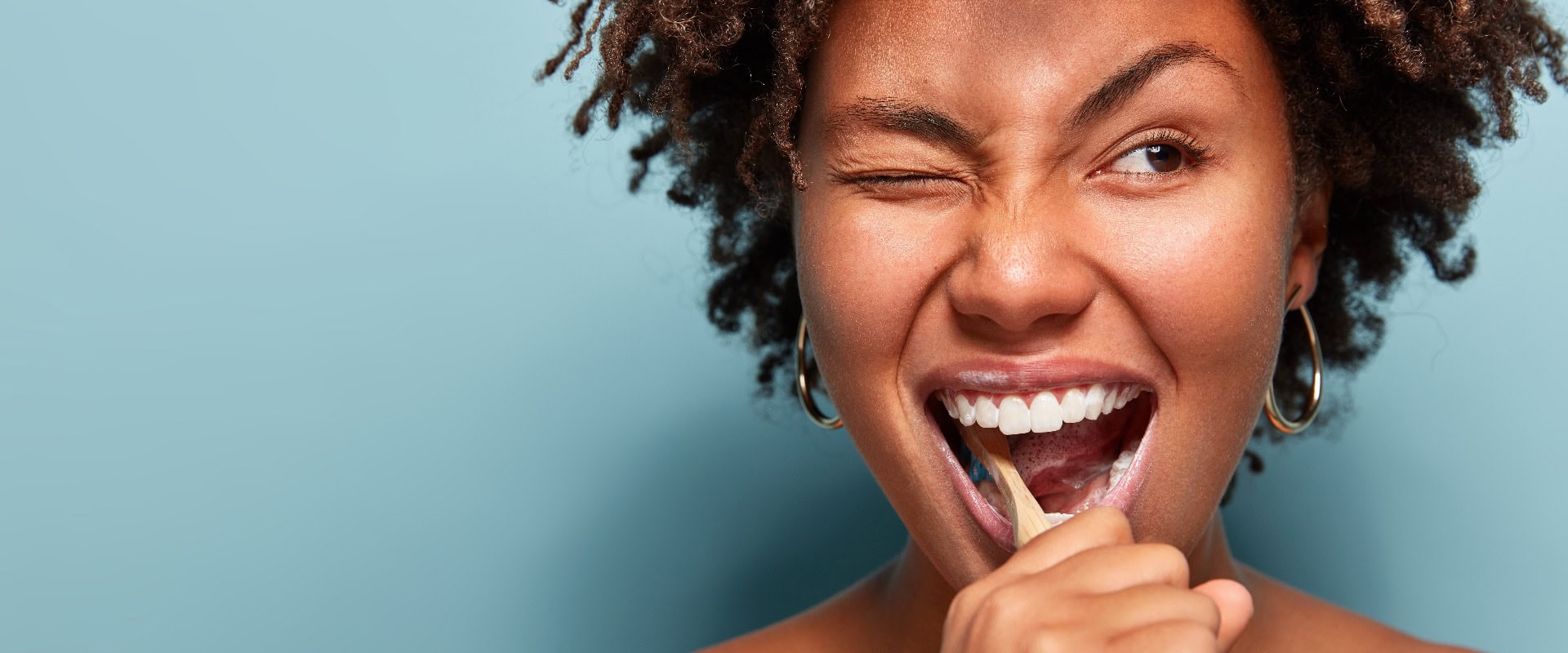 Nahaufnahme einer fröhlichen, lustigen, dunkelhäutigen jungen Frau mit Afro-Haar, die sich aktiv mit der Zahnbürste die Zähne putzt, den Mund weit öffnet, mit den Augen zwinkert, die nackten Schultern zeigt und sich um die Mundhygiene kümmert