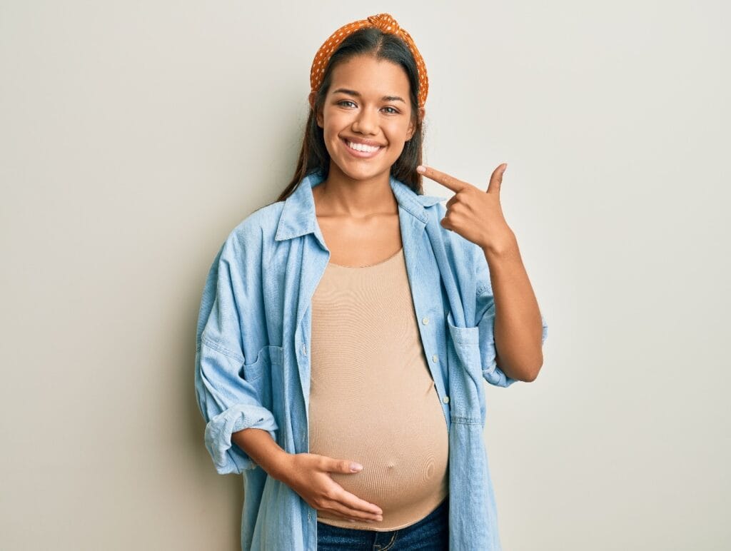 Schöne hispanische Frau, die ein Baby erwartet, berührt den schwangeren Bauch, lächelt fröhlich und zeigt mit den Fingern Zähne und Mund. Zahngesundheit Konzept.